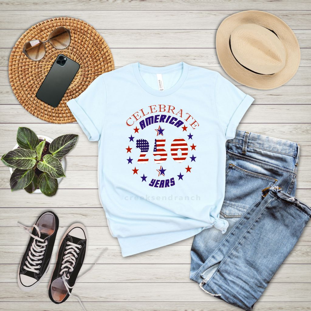 Flat lay of a light blue Celebrate America T-shirt with stars and stripes, denim jeans, straw hat, sneakers, sunglasses, and phone on wood surface.