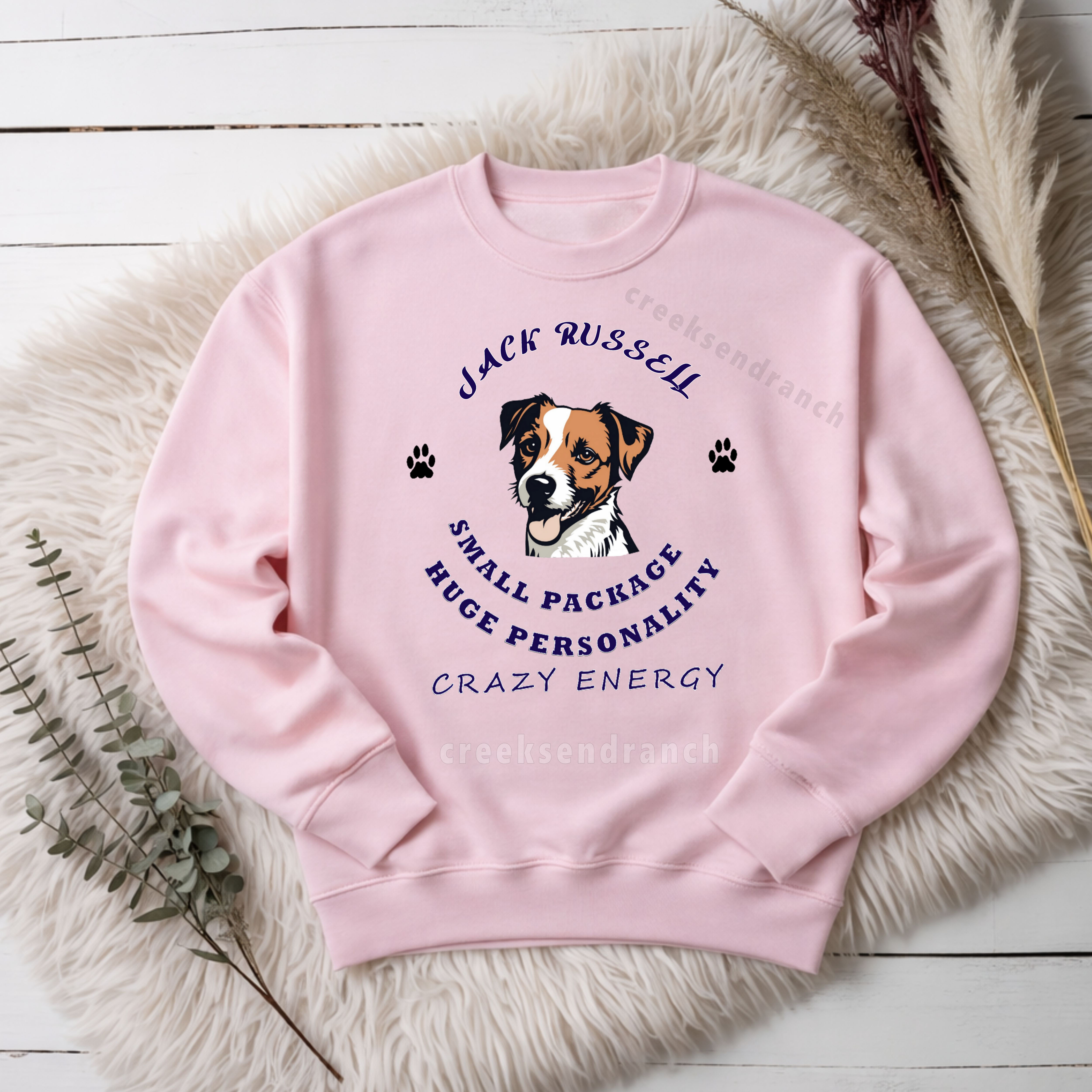 Pink sweatshirt with a Jack Russell dog graphic and curved navy text, placed on a white wooden floor with a fluffy rug nearby.