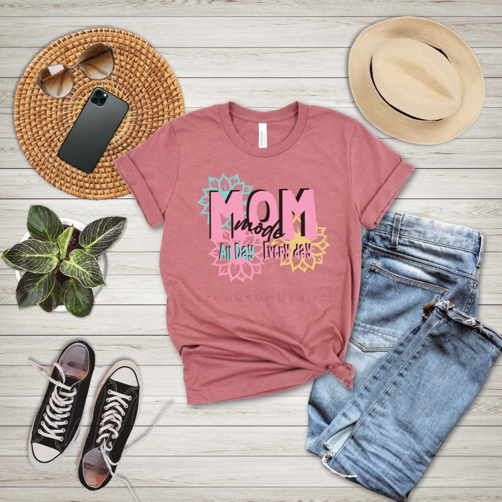 Overhead view of a pink graphic T-shirt with 'Mom' design, denim shorts, black sneakers, a straw hat, sunglasses, and a woven mat on a light wooden surface.